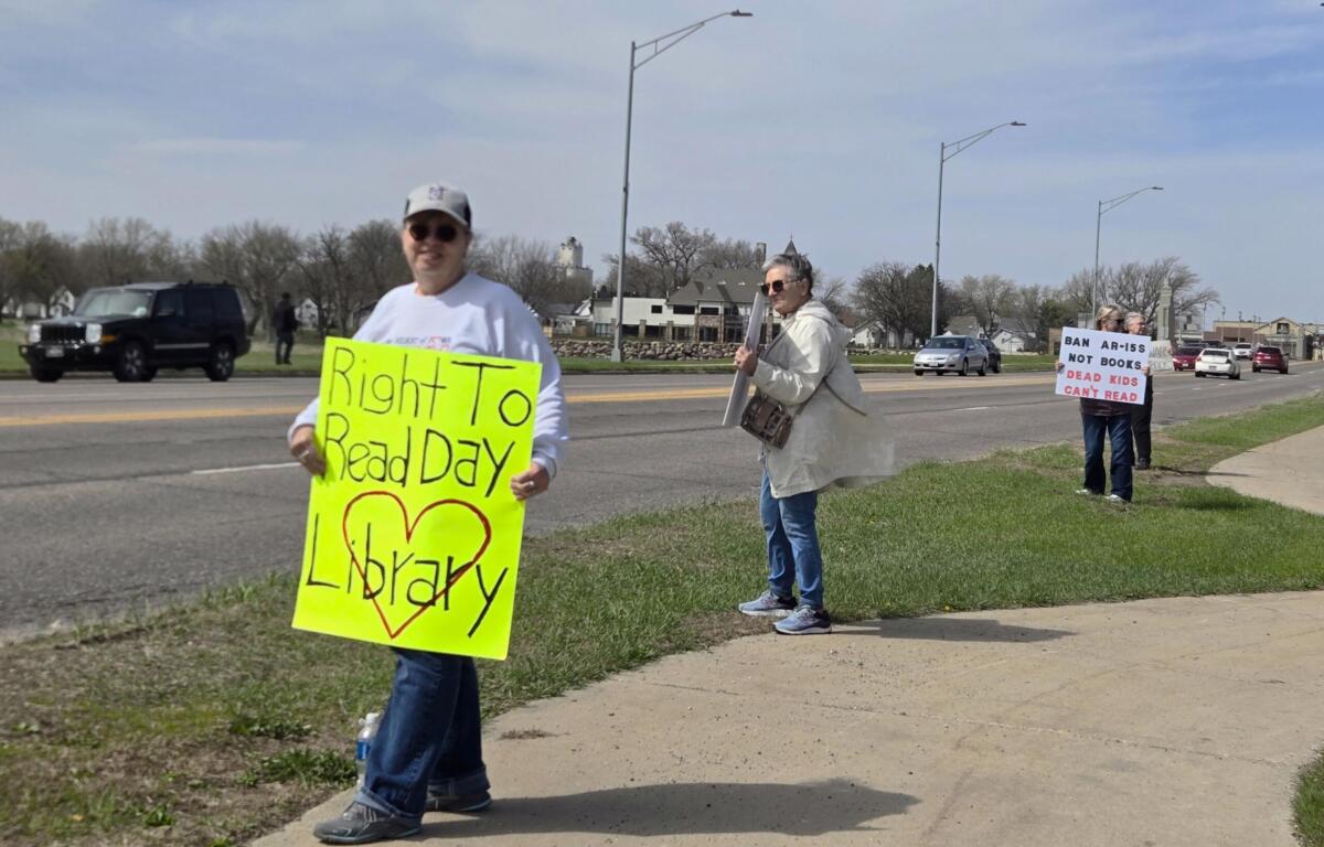 A roadside protest with people holding bright signs; foreground signer holds a fluorescent yellow poster about the 'Right To Read Day'.