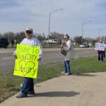 A roadside protest with people holding bright signs; foreground signer holds a fluorescent yellow poster about the 'Right To Read Day'.