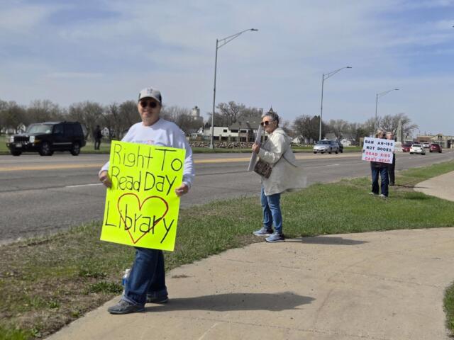 A roadside protest with people holding bright signs; foreground signer holds a fluorescent yellow poster about the 'Right To Read Day'.