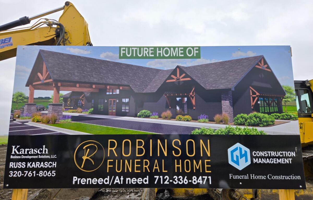 Construction sign showing a rendering of the future Robinson Funeral Home, with a yellow excavator arm in the background and contact information on the board.
