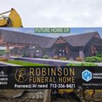 Construction sign showing a rendering of the future Robinson Funeral Home, with a yellow excavator arm in the background and contact information on the board.