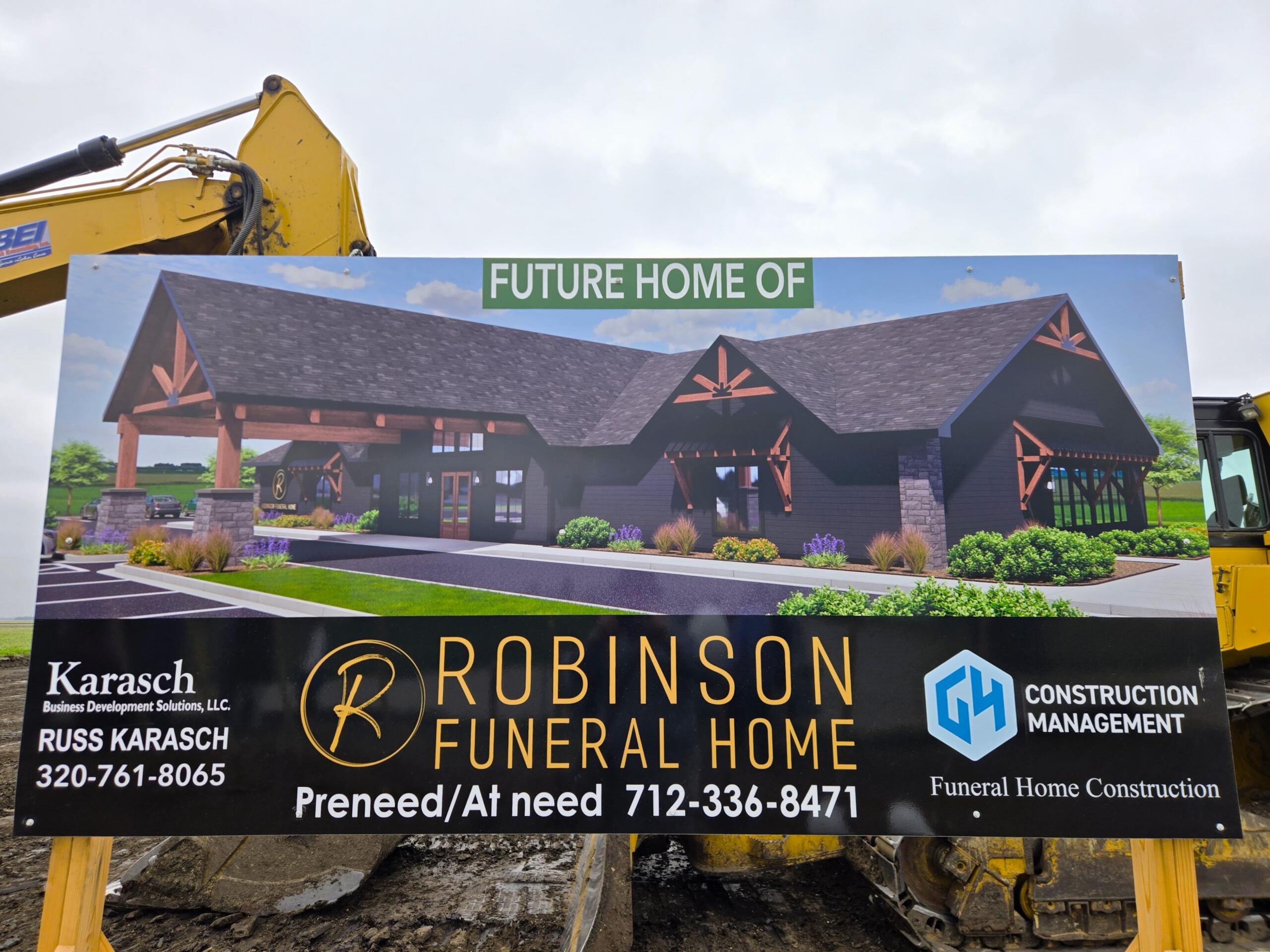 Construction sign showing a rendering of the future Robinson Funeral Home, with a yellow excavator arm in the background and contact information on the board.
