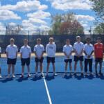 Group of young tennis players standing shoulder to shoulder on a blue outdoor court in front of a net, wearing white shirts and shorts.