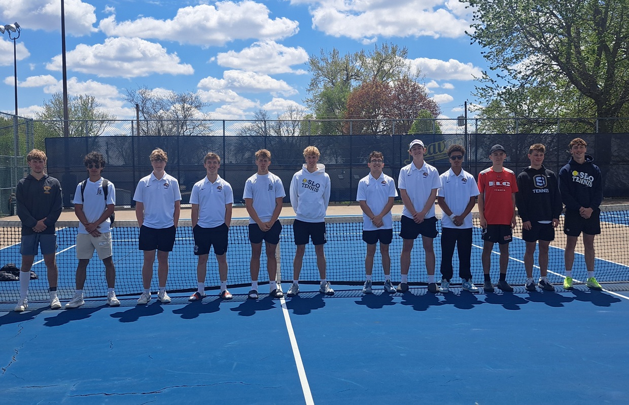 Group of young tennis players standing shoulder to shoulder on a blue outdoor court in front of a net, wearing white shirts and shorts.