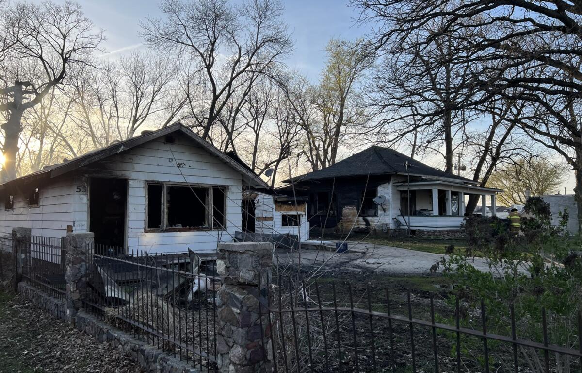 Two neighboring houses with severe fire damage: charred exteriors and burnt windows, debris in the yards, a firefighter in turnout gear near the right side, with leafless trees behind.