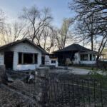 Two neighboring houses with severe fire damage: charred exteriors and burnt windows, debris in the yards, a firefighter in turnout gear near the right side, with leafless trees behind.