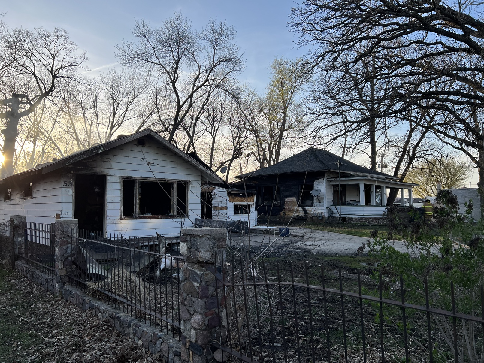 Two neighboring houses with severe fire damage: charred exteriors and burnt windows, debris in the yards, a firefighter in turnout gear near the right side, with leafless trees behind.