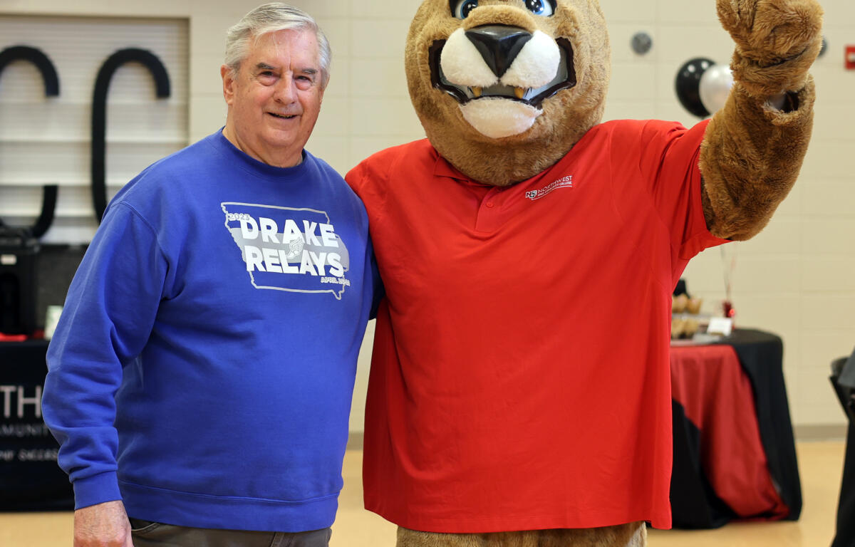Older man in a blue Drake Relays T-shirt posing beside a large brown lion mascot in a red shirt at an indoor event.