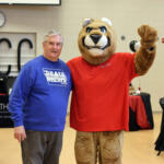 Older man in a blue Drake Relays T-shirt posing beside a large brown lion mascot in a red shirt at an indoor event.