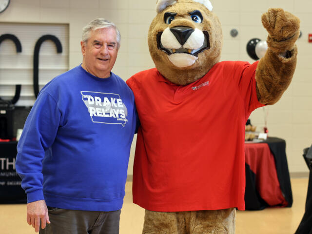Older man in a blue Drake Relays T-shirt posing beside a large brown lion mascot in a red shirt at an indoor event.