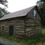 Old log cabin with a pitched shingle roof in a grassy park setting, daytime light fading to dark.