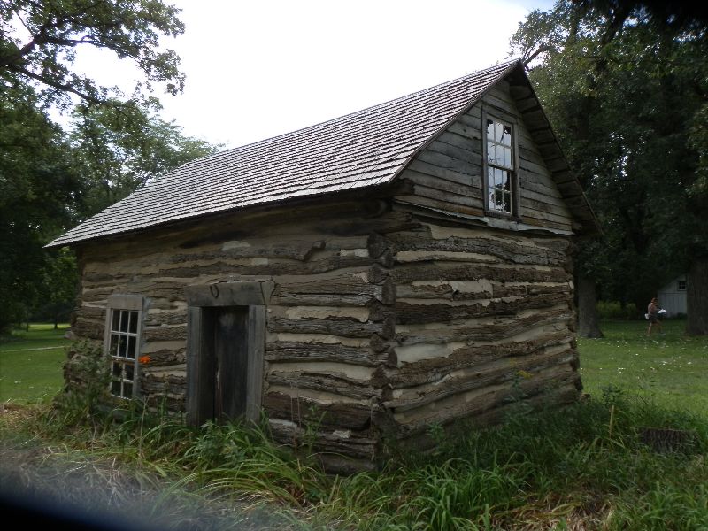 Old log cabin with a pitched shingle roof in a grassy park setting, daytime light fading to dark.
