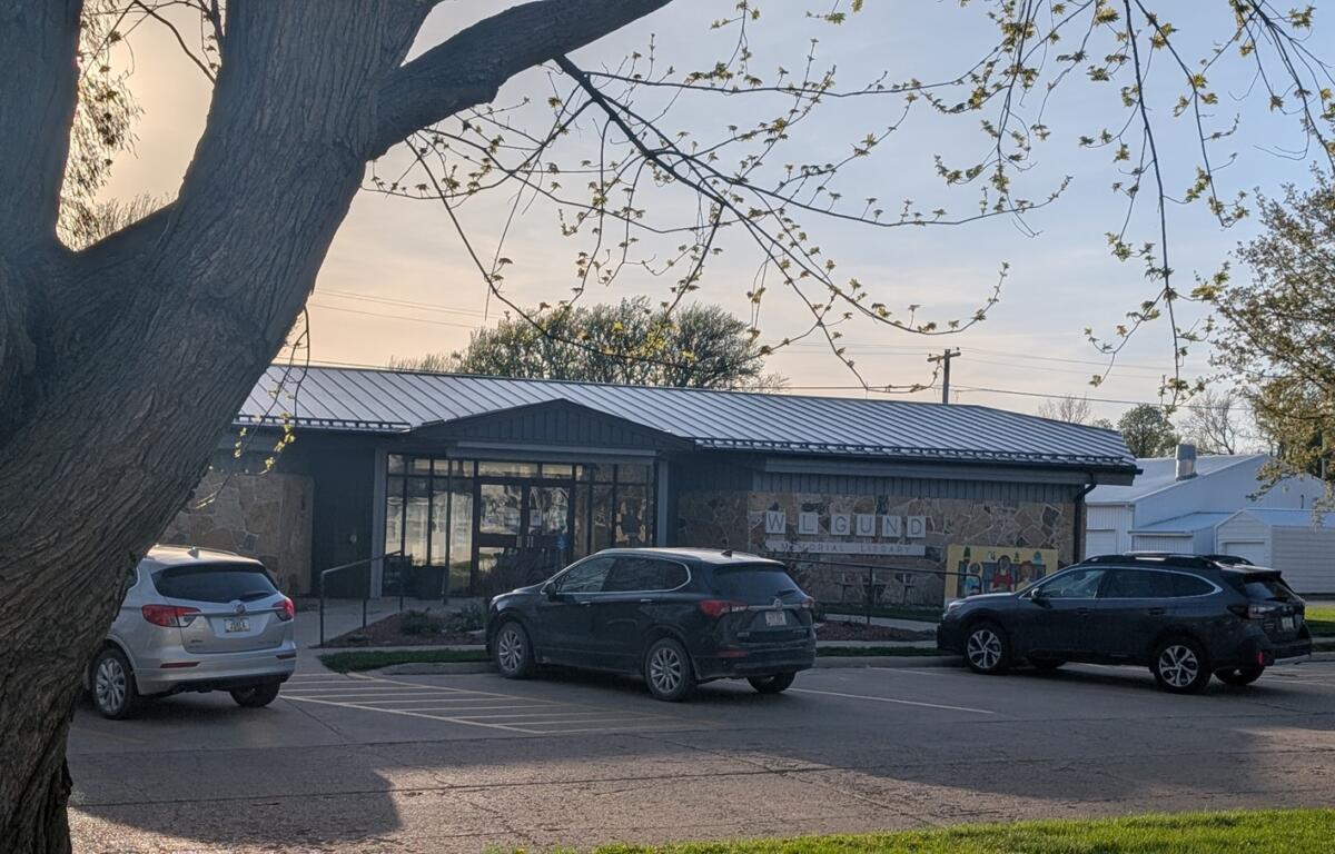 Exterior view of a memorial library with a stone wall and metal roof, parked cars in front, and a large tree framing the scene.