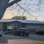 Exterior view of a memorial library with a stone wall and metal roof, parked cars in front, and a large tree framing the scene.