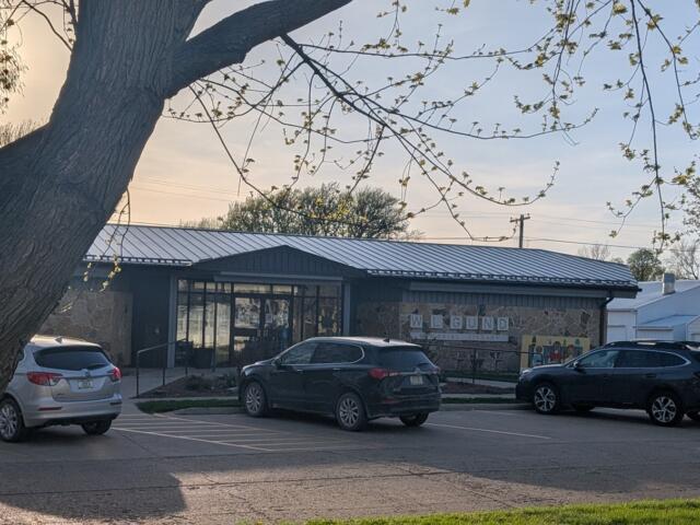 Exterior view of a memorial library with a stone wall and metal roof, parked cars in front, and a large tree framing the scene.