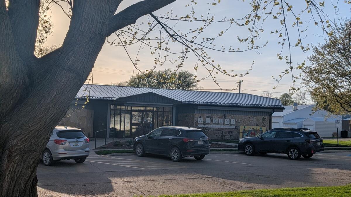 Exterior view of a memorial library with a stone wall and metal roof, parked cars in front, and a large tree framing the scene.