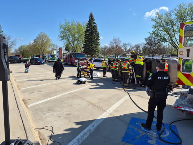 Emergency responders and a police officer surround an overturned vehicle during a rescue at a parking lot.