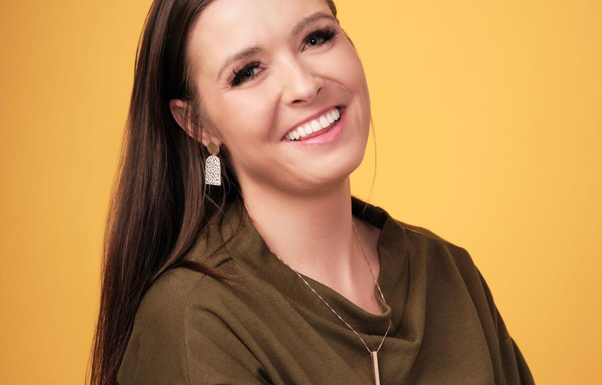 Smiling woman with long dark hair wearing an olive-green top and a pendant against a solid yellow background.