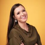 Smiling woman with long dark hair wearing an olive-green top and a pendant against a solid yellow background.