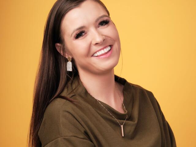 Smiling woman with long dark hair wearing an olive-green top and a pendant against a solid yellow background.
