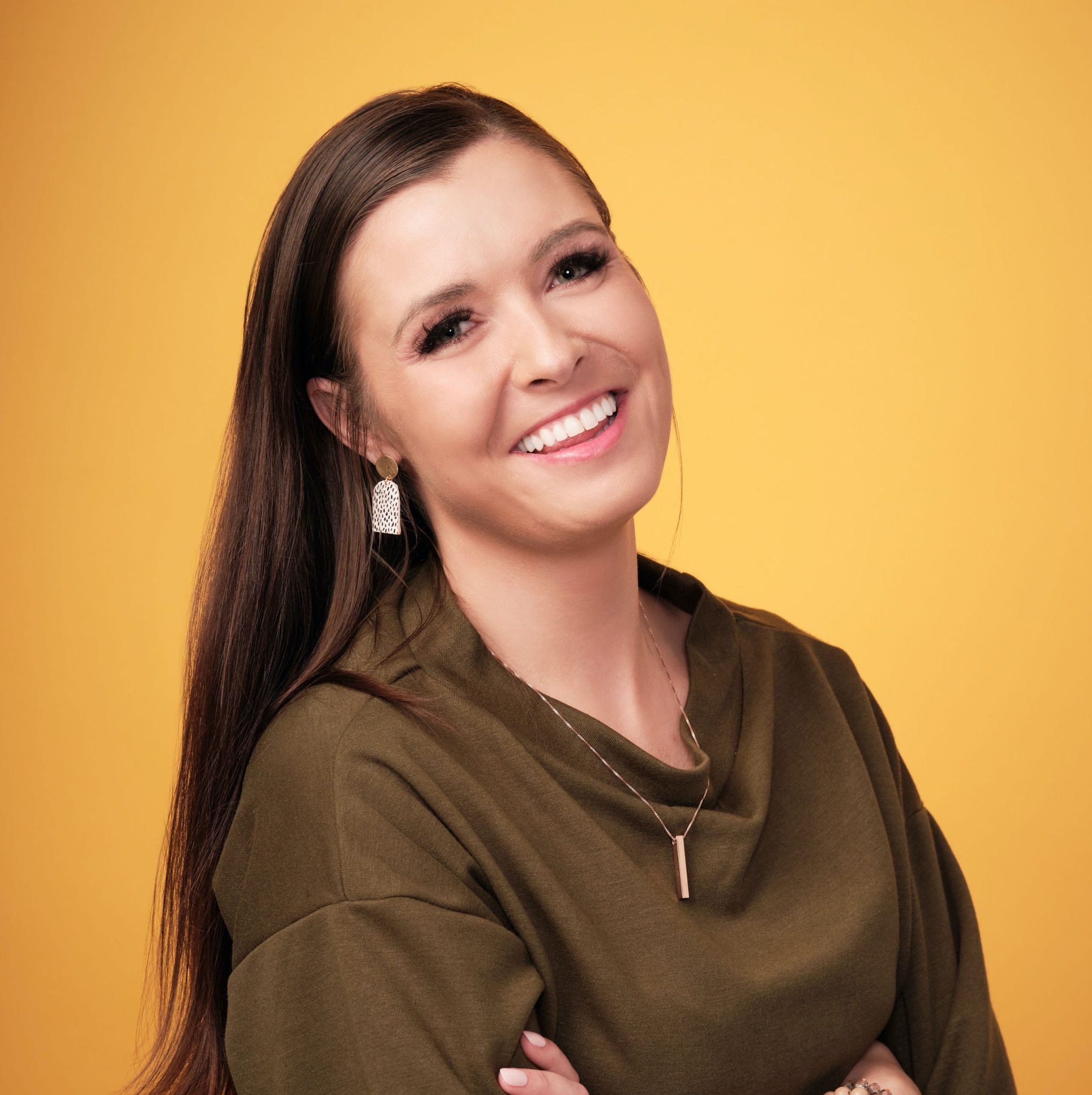 Smiling woman with long dark hair wearing an olive-green top and a pendant against a solid yellow background.