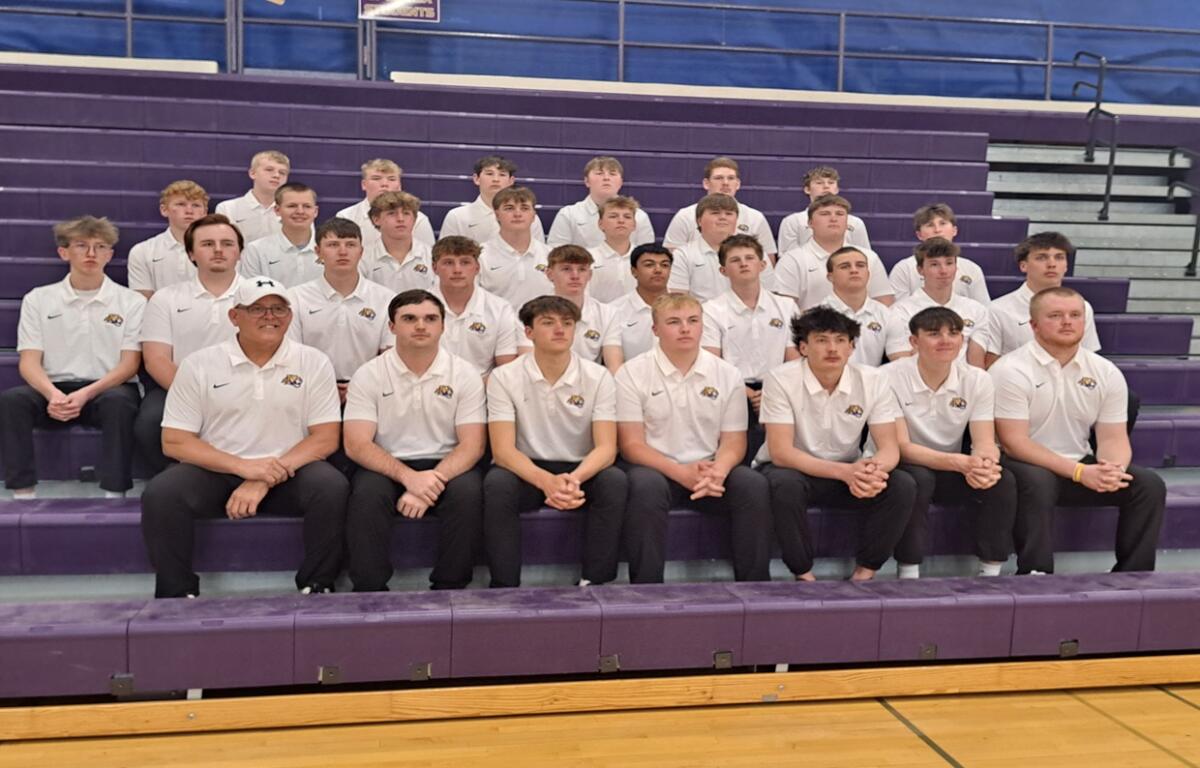 Group of about 25 male students in white polo shirts and black pants posing on purple gym bleachers for a team photo.