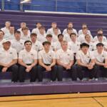Group of about 25 male students in white polo shirts and black pants posing on purple gym bleachers for a team photo.