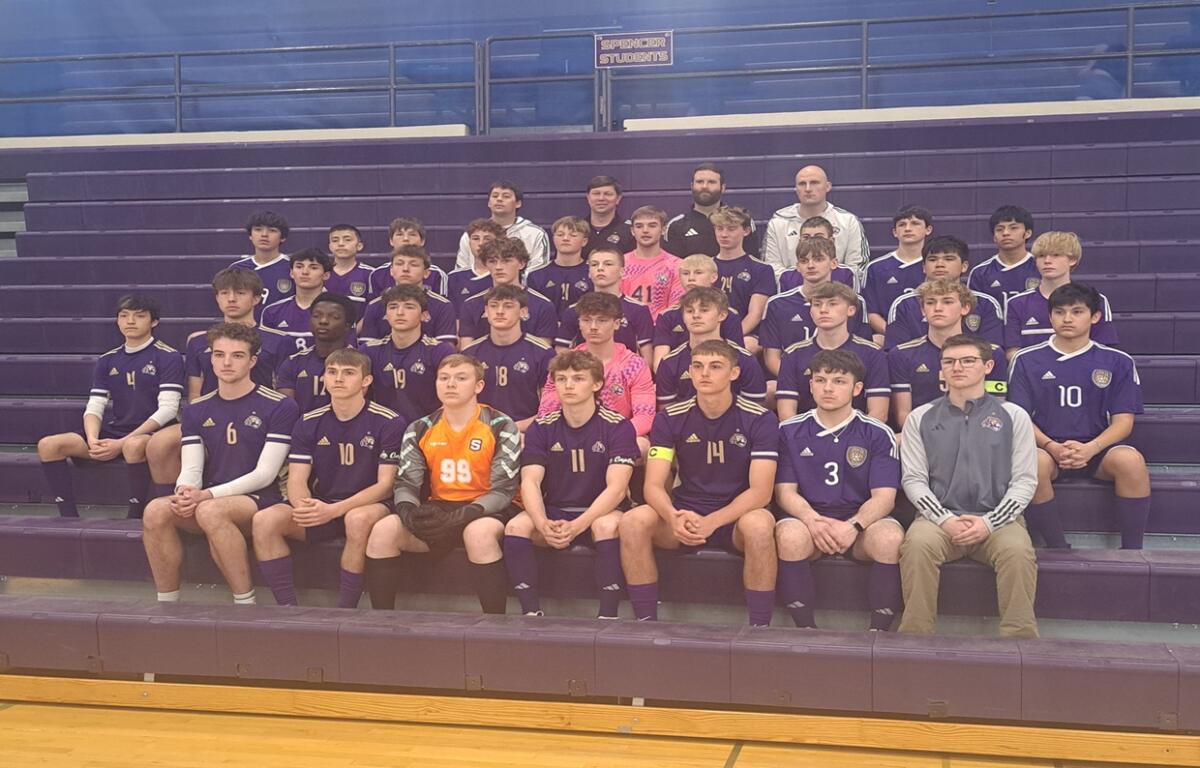Group photo of a boys' soccer team in purple uniforms with numbers, sitting on gym bleachers with two coaches in the back row.