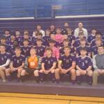 Group photo of a boys' soccer team in purple uniforms with numbers, sitting on gym bleachers with two coaches in the back row.