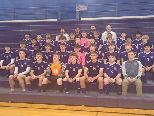 Group photo of a boys' soccer team in purple uniforms with numbers, sitting on gym bleachers with two coaches in the back row.