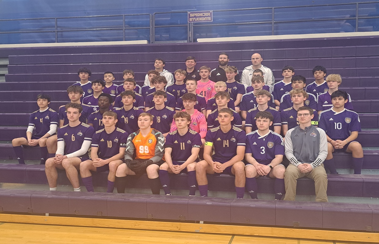 Group photo of a boys' soccer team in purple uniforms with numbers, sitting on gym bleachers with two coaches in the back row.