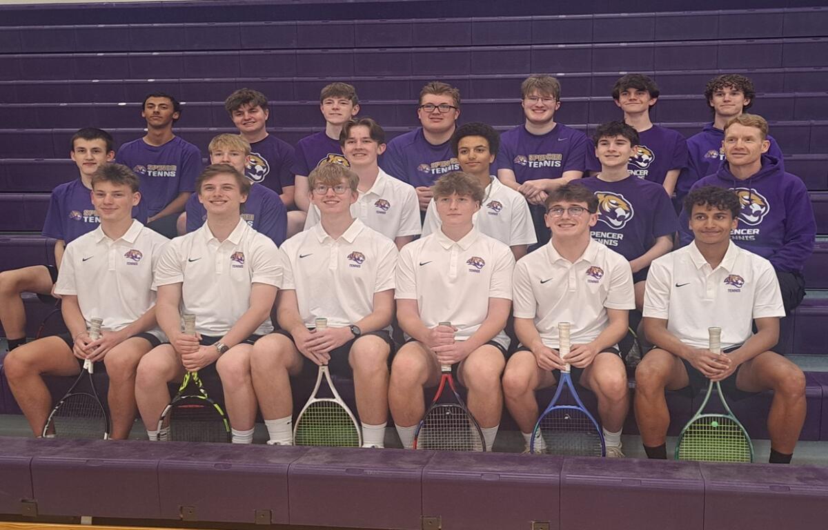 Group of tennis players posing in a gym bleacher photo; front row in white polo shirts, back row in purple shirts, each holding rackets.