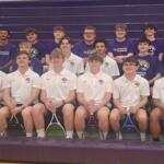 Group of tennis players posing in a gym bleacher photo; front row in white polo shirts, back row in purple shirts, each holding rackets.