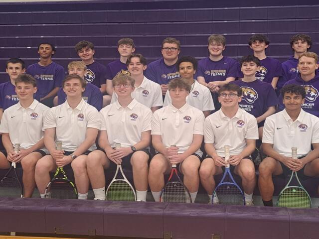 Group of tennis players posing in a gym bleacher photo; front row in white polo shirts, back row in purple shirts, each holding rackets.