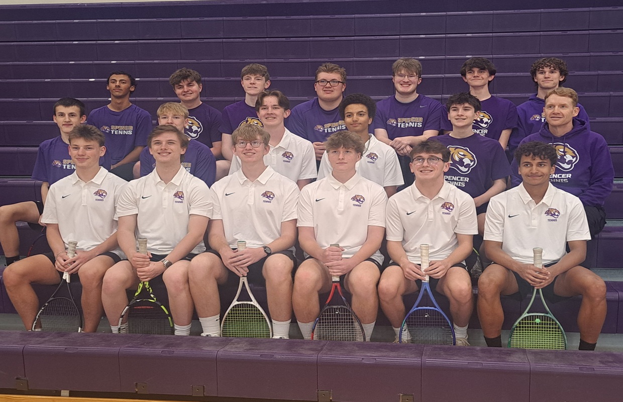 Group of tennis players posing in a gym bleacher photo; front row in white polo shirts, back row in purple shirts, each holding rackets.