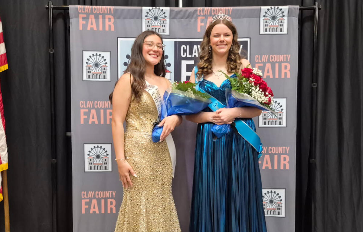 Two young women in formal gowns pose with bouquets at a Clay County Fair backdrop, one wearing a gold sequin dress and sash, the other in a blue gown with a tiara.