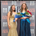 Two young women in formal gowns pose with bouquets at a Clay County Fair backdrop, one wearing a gold sequin dress and sash, the other in a blue gown with a tiara.