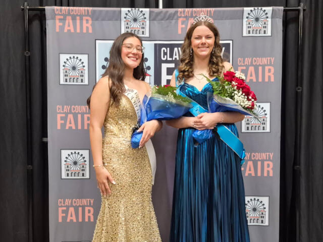Two young women in formal gowns pose with bouquets at a Clay County Fair backdrop, one wearing a gold sequin dress and sash, the other in a blue gown with a tiara.