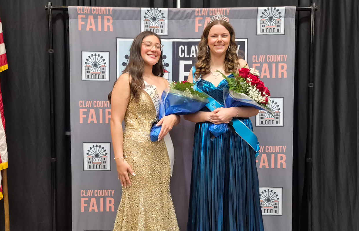 Two young women in formal gowns pose with bouquets at a Clay County Fair backdrop, one wearing a gold sequin dress and sash, the other in a blue gown with a tiara.
