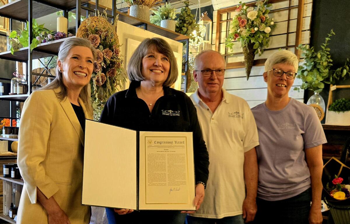 Four adults smile for a photo indoors, one woman presenting a framed certificate as others stand beside her in a floral shop setting.