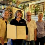 Four adults smile for a photo indoors, one woman presenting a framed certificate as others stand beside her in a floral shop setting.