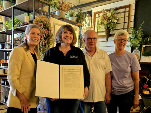 Four adults smile for a photo indoors, one woman presenting a framed certificate as others stand beside her in a floral shop setting.