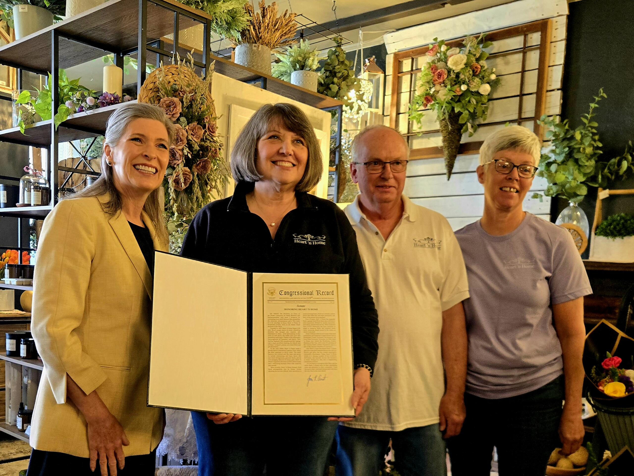 Four adults smile for a photo indoors, one woman presenting a framed certificate as others stand beside her in a floral shop setting.