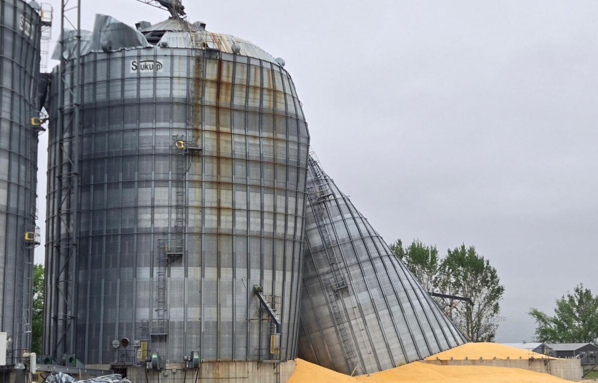 Tall corrugated metal grain silos with rust streaks and a collapsed top section against a gray sky; orange sand piles at the base suggest industrial site.