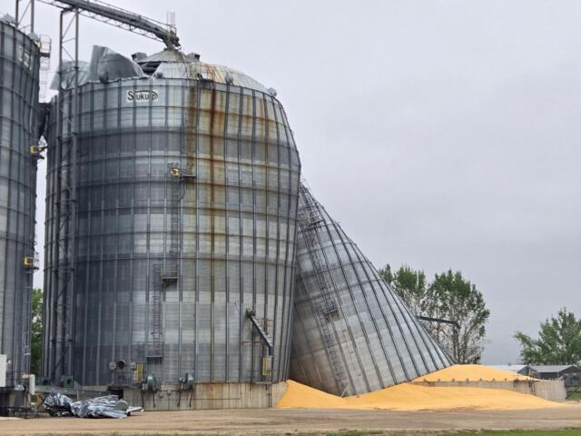 Tall corrugated metal grain silos with rust streaks and a collapsed top section against a gray sky; orange sand piles at the base suggest industrial site.