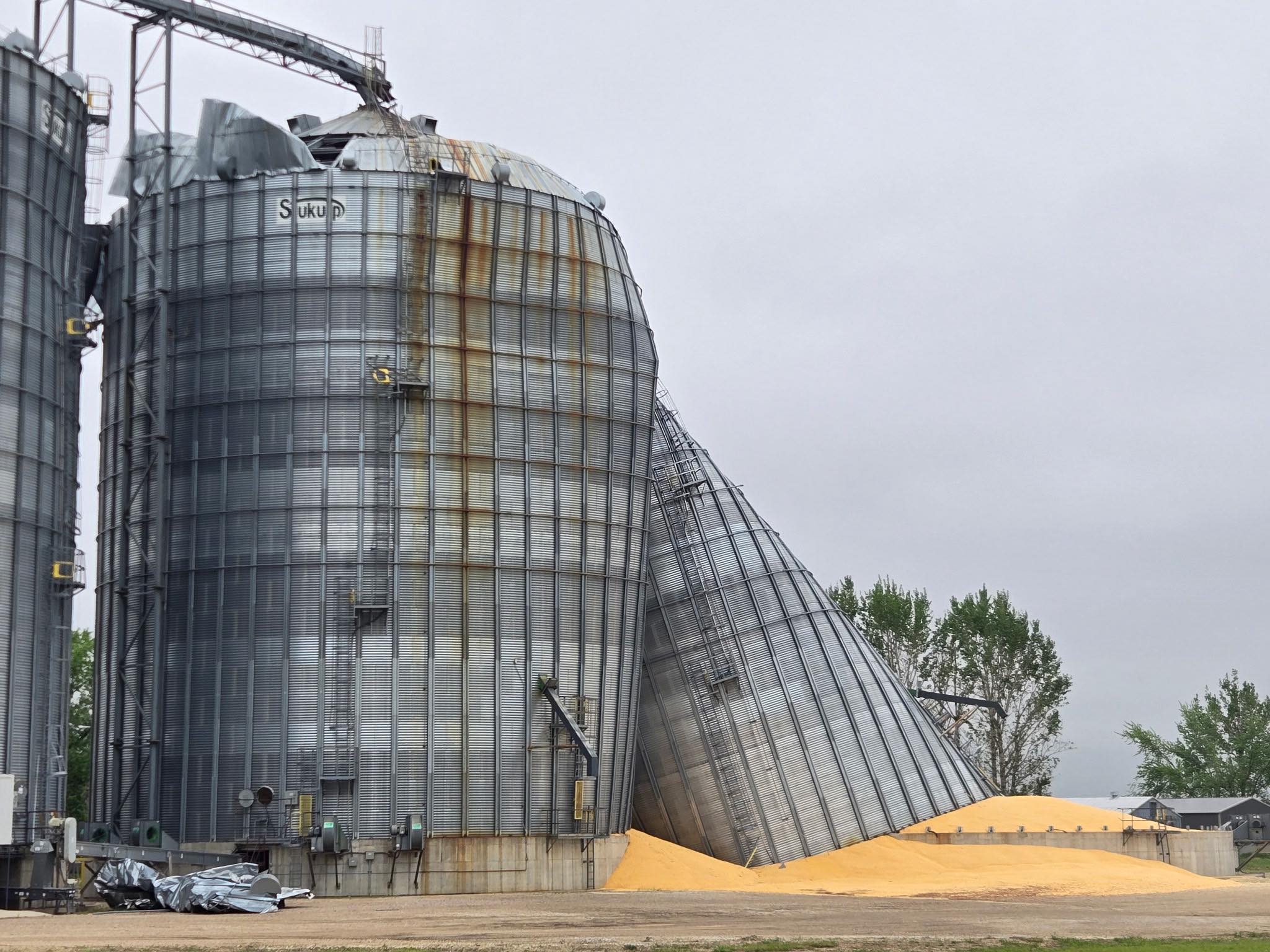 Tall corrugated metal grain silos with rust streaks and a collapsed top section against a gray sky; orange sand piles at the base suggest industrial site.