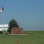 Stone sign reading 'MARCUS' with 'FOUNDED IN' text, flanked by flags on the left and a white industrial building in the background on a green lawn.
