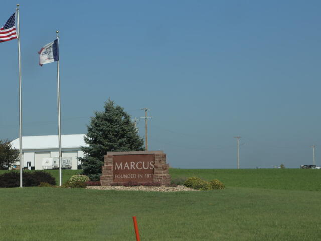 Stone sign reading 'MARCUS' with 'FOUNDED IN' text, flanked by flags on the left and a white industrial building in the background on a green lawn.