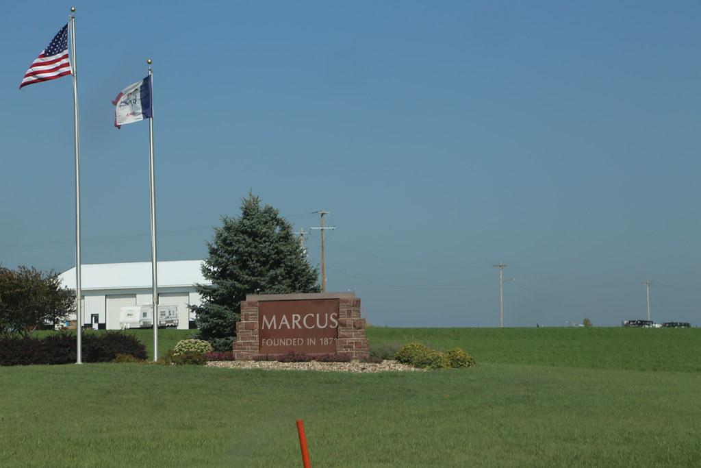 Stone sign reading 'MARCUS' with 'FOUNDED IN' text, flanked by flags on the left and a white industrial building in the background on a green lawn.