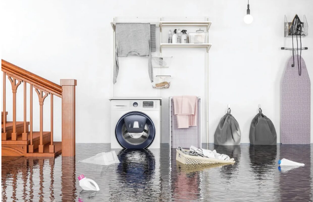 Laundry room flooded with water on the floor; a front-loading washer sits beside a drying rack and ironing board visible in the background.